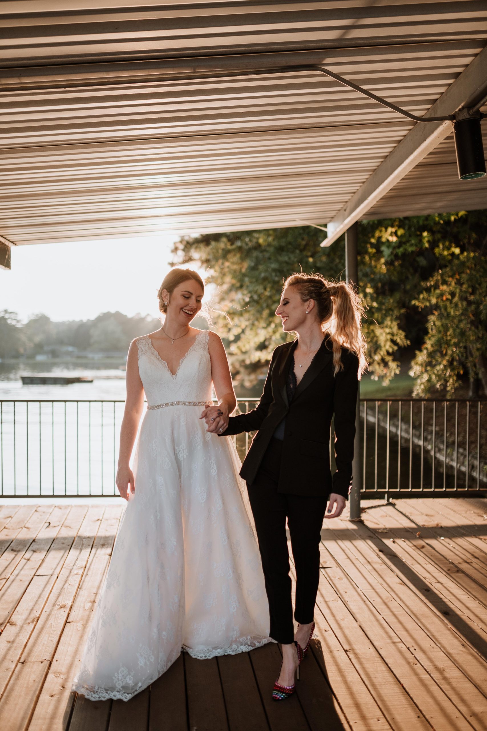 Lesbian newlywed couple walking together along a dock at sunset.
