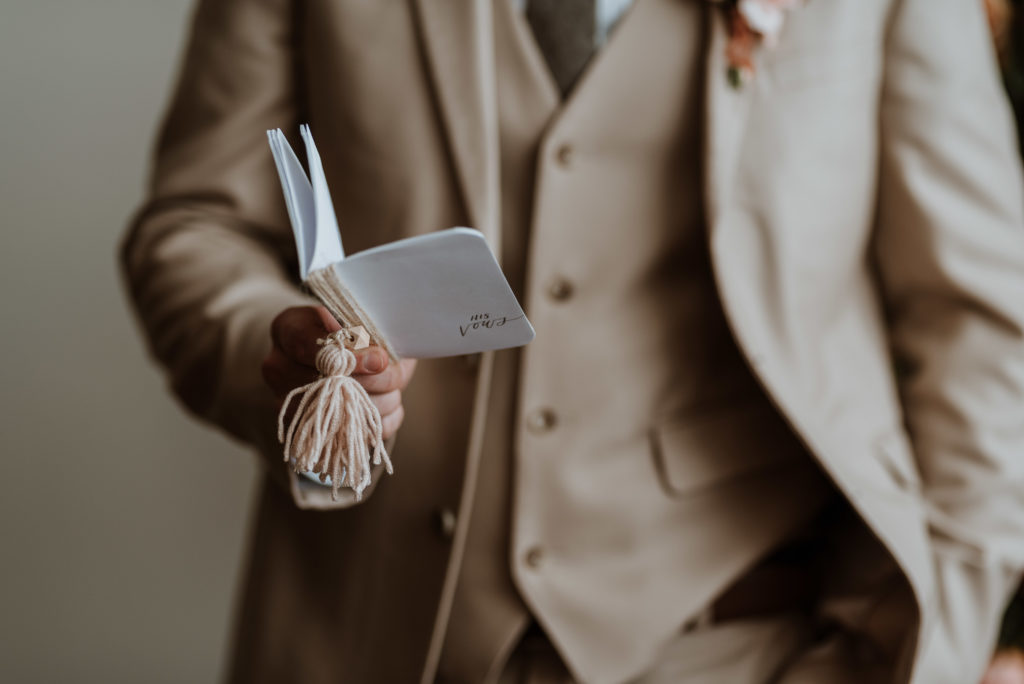 Groom in tan suit holding small vow book as he reads his written vows to his partner.